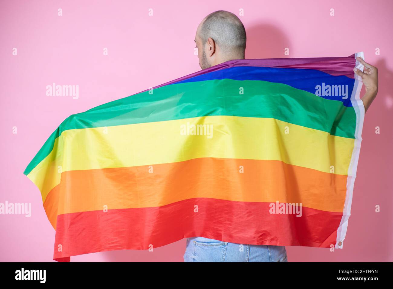Rückansicht eines jungen, hübschen, kahlen Mannes aus Mexiko, der mit einer LGBT-Flagge vor einem rosa Hintergrund posiert Stockfoto