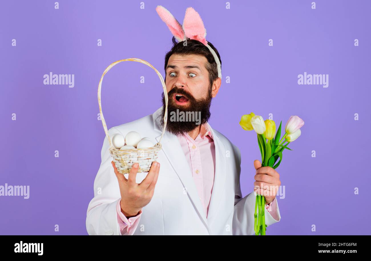 Ostern. Kaninchenmann in Hasenohren mit Blume. Bärtiger Rüde in Anzug hält Korb mit Eiern. Stockfoto