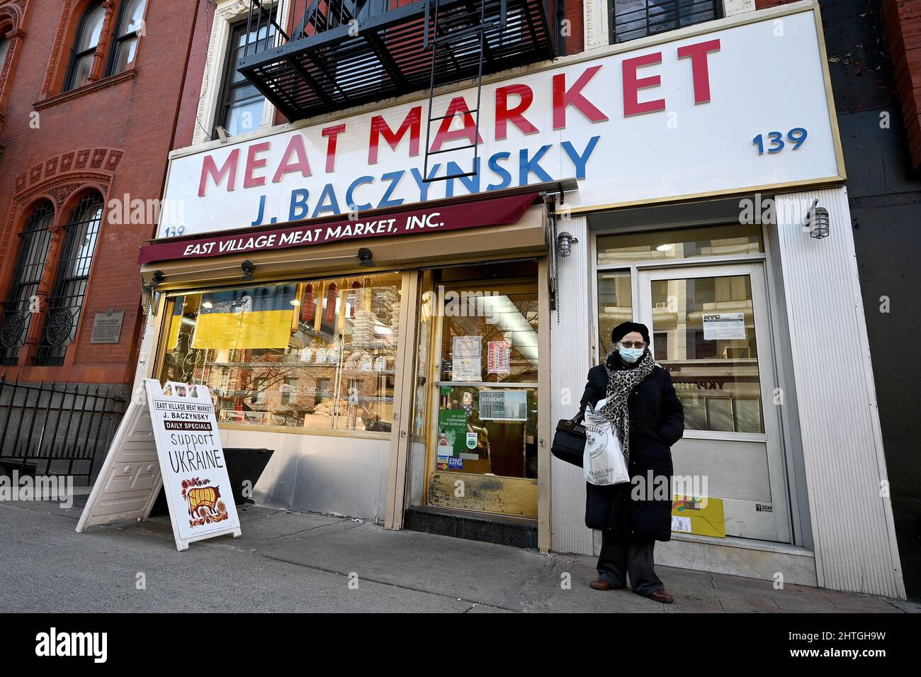 New York, USA. 28.. Februar 2022. Eine Frau steht vor dem J. Baczynsky Meat Market, wo eine ukrainische Flagge und ein Schild ihre Unterstützung für die Ukraine nach dem Einmarsch von Russland, New York, NY, 28. Februar 2022, markieren. Der russische Präsident Wladimir Putin begann in den frühen Morgenstunden des 24. Februar mit der Invasion in die benachbarte Ukraine. (Foto von Anthony Behar/Sipa USA) Quelle: SIPA USA/Alamy Live News Stockfoto