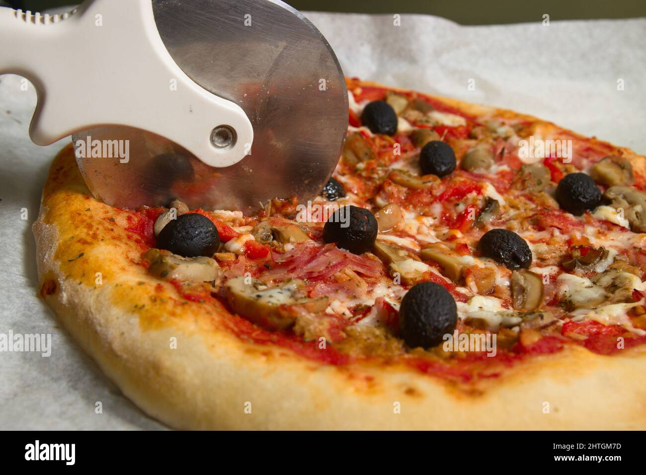 Eine hausgemachte Pizza mit Tomatensauce, Pilzen und schwarzen Oliven, die mit einem Cutter auf dem Ofenblech geschnitten werden Stockfoto