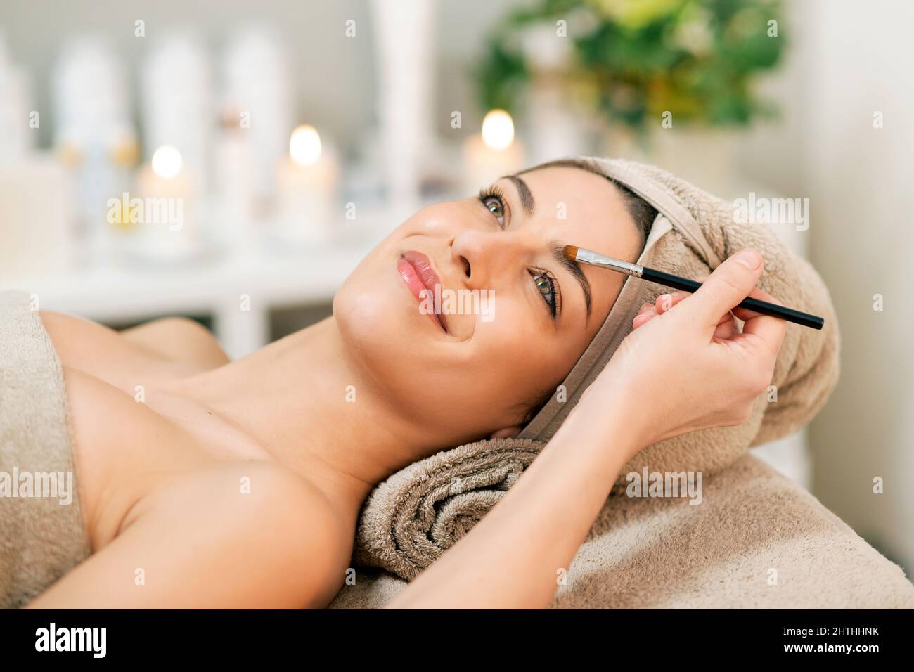 Crop weibliche Beauty Master mit Pinsel Anwendung Augenbrauen Schatten auf Gesicht der lächelnden Frau in Handtuch liegend auf der Couch im Salon während der entspannenden Therapie-Sitzung Stockfoto