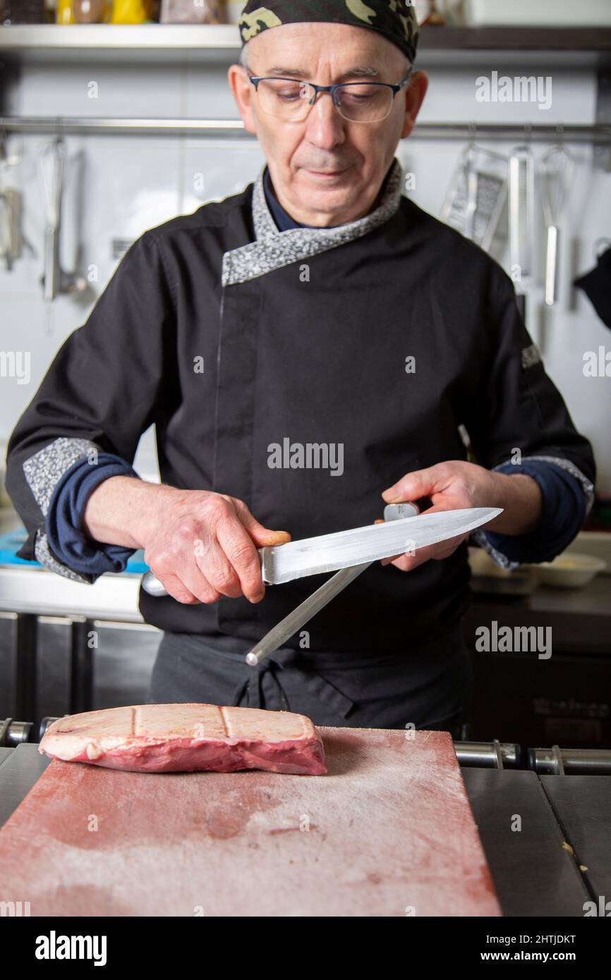 Reifer männlicher Koch in einheitlicher Schärfklinge aus Messer mit Honstahl, während er sich für das Kochen von Rindfleisch in der Küche des Restaurants vorbereitet Stockfoto