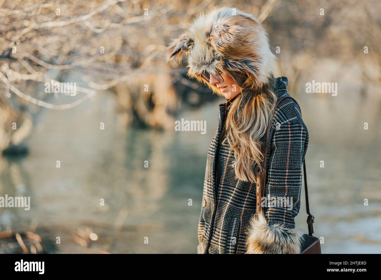 Seitenansicht modische Frau in Sonnenbrillen tragen Tasse und Mantel mit Stulpen aus Fuchspelz auf verschwommenem Hintergrund von See Wasser nach unten schauen Stockfoto