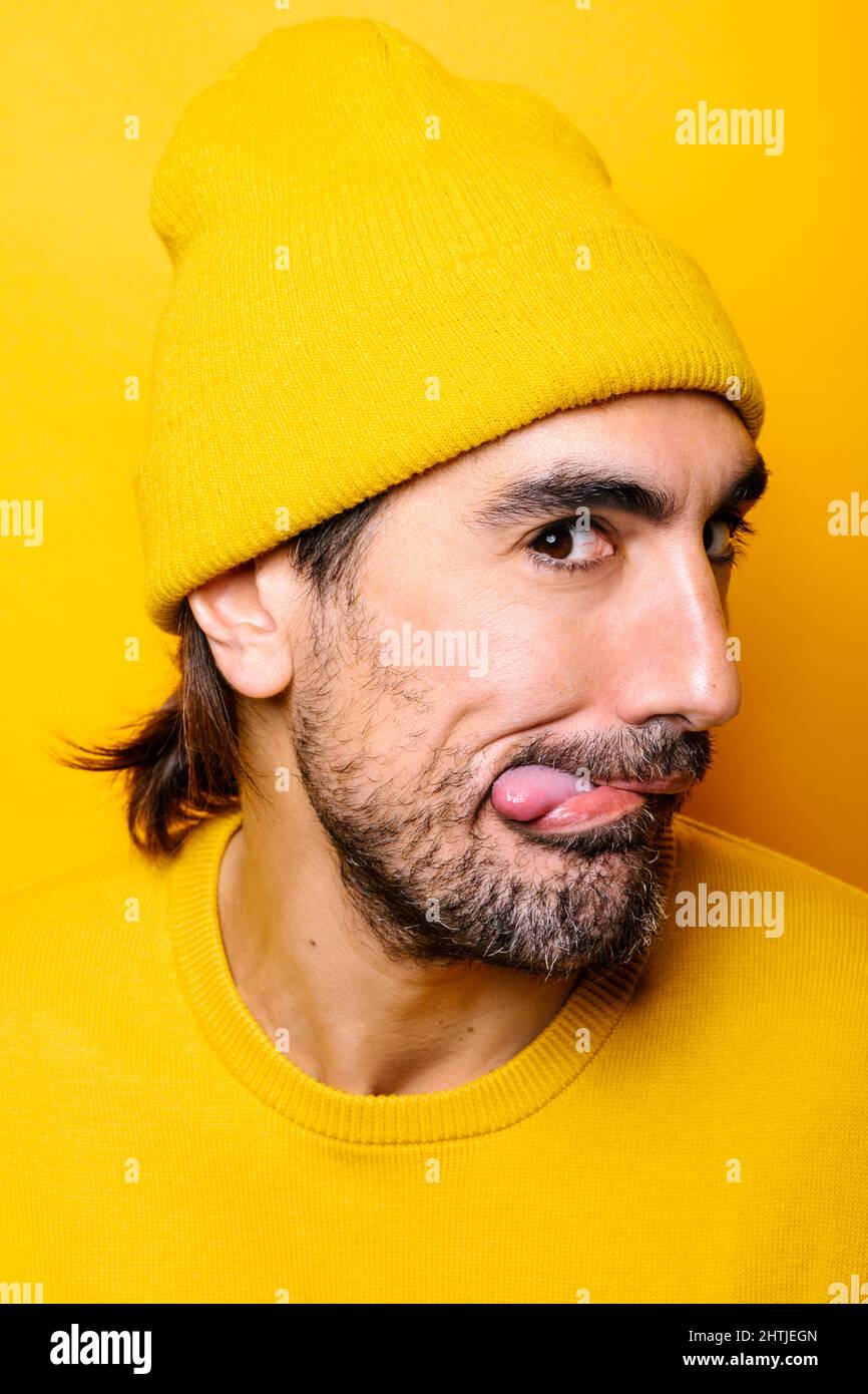 Sorgloser bärtiger Mann mit dunklem Haar im Hut, der Zunge zeigt und im hellen Studio vor gelbem Hintergrund auf die Kamera schaut Stockfoto