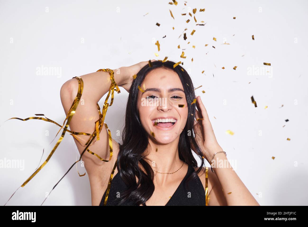 Positive junge Frau, die mit einem Lächeln auf die Kamera blickt, während sie während der Feiertagsfeier im Studio unter herabfallenden Konfetti auf weißem Hintergrund steht Stockfoto