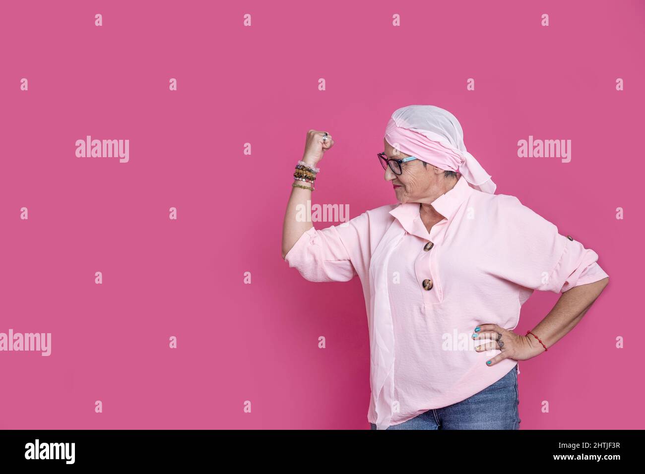 Positive reife Frau mit Krebs trägt Kopftuch und zeigt Bizep, während in der Nähe von rosa Hintergrund in hellen Studio stehen Stockfoto