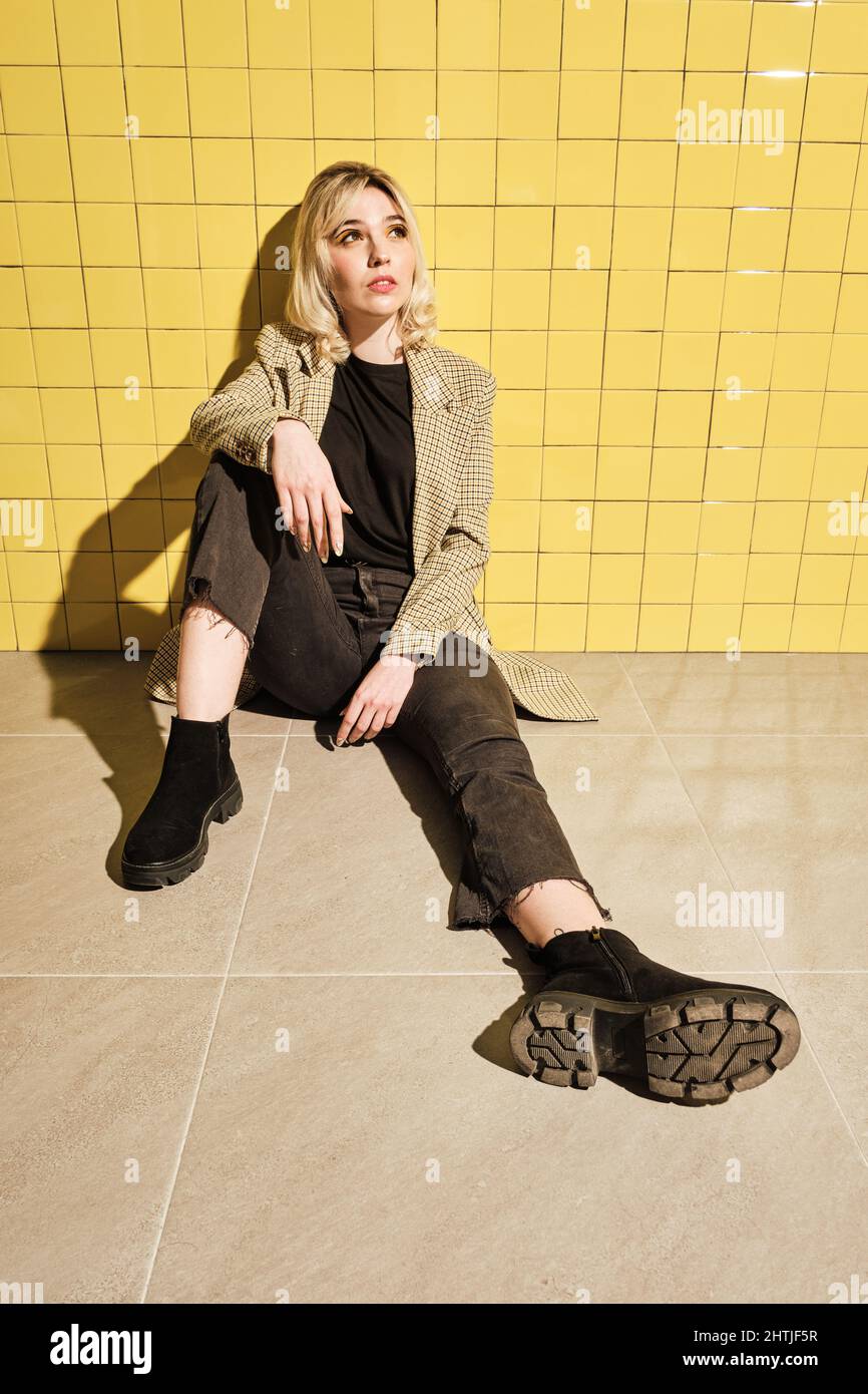 Ganzer Körper der jungen Frau in stilvollem Outfit weg schauen, während sitzen in der Nähe gelb geflieste Wand Stockfoto
