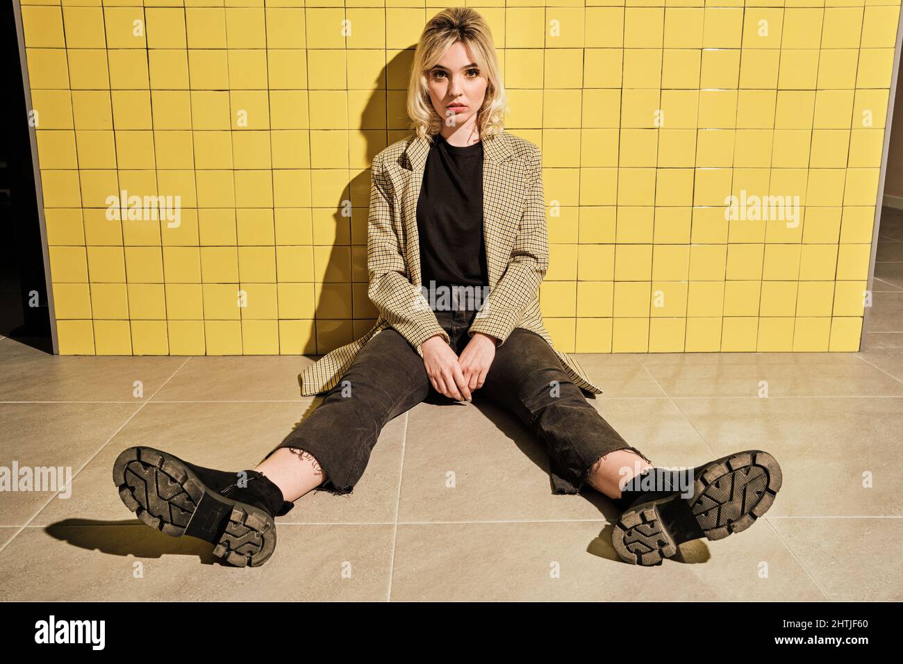 Ganzer Körper von jungen Weibchen in stilvollem Outfit Blick auf die Kamera, während sie in der Nähe der gelb gefliesten Wand sitzen Stockfoto