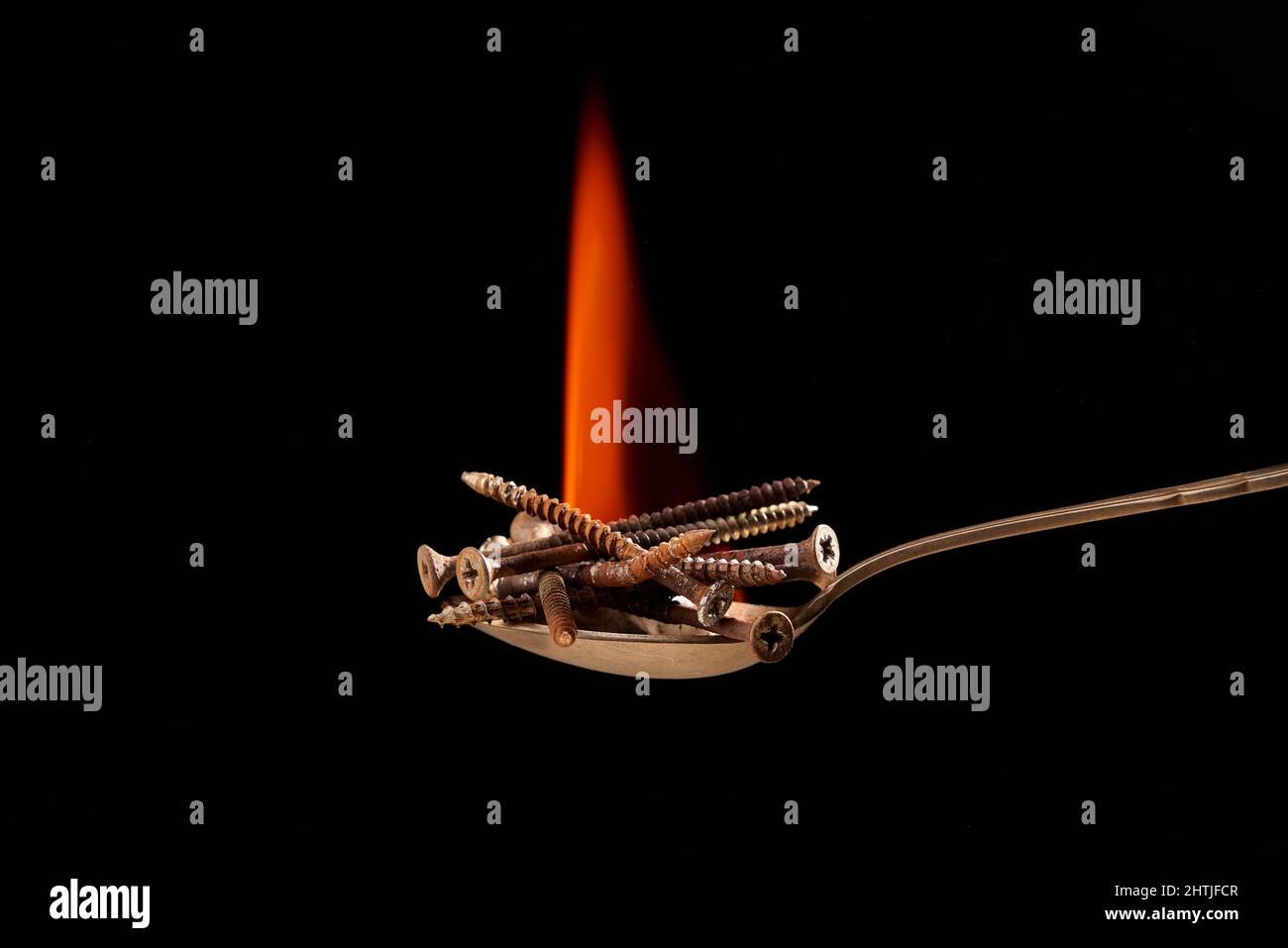 Kleiner Haufen schwerer ungesunder Metallschrauben, die Junk Food darstellen, auf Löffel mit oranger Flamme auf schwarzem Hintergrund im Studio platziert Stockfoto