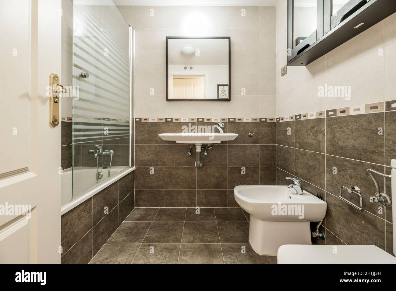 Großes Badezimmer mit separater Dusche, weißen Porzellantoiletten, Fliesenverkleidung, grauem Fliesen- und braunem Spiegel und schwarzen Schränken Stockfoto