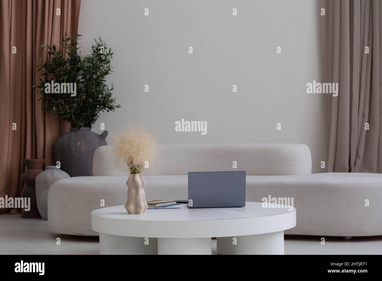 Komposition aus weißem runden Tisch mit Laptop und handgefertigter Vase gegen bequemes Sofa und beigefarbene Vorhänge an der Wand Stockfoto