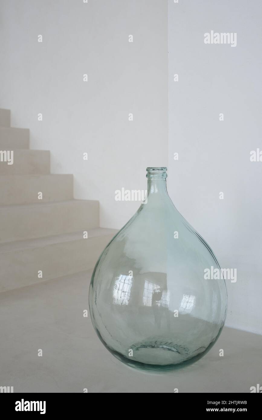 Große tropfenförmige Glasvase, die auf dem Boden des modernen, stilvollen Wohnzimmers in weißer Farbe gegen Treppe und Wand steht Stockfoto