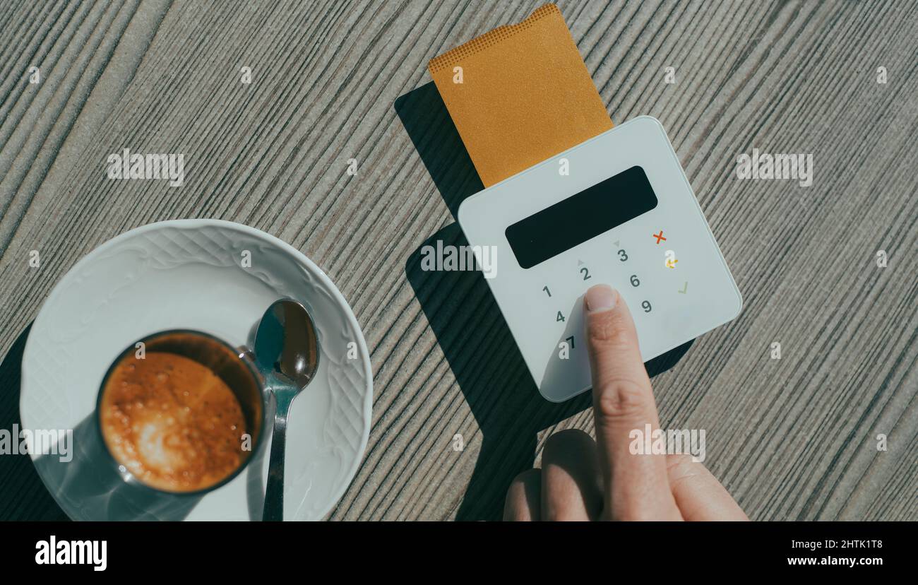 Blick aus der Perspektive eines jungen Mannes auf einen Tisch in einem Straßencafé, der den PIN-Code seiner Kreditkarte in das Yepad eines drahtlosen Zahlungsterminals einfügt Stockfoto