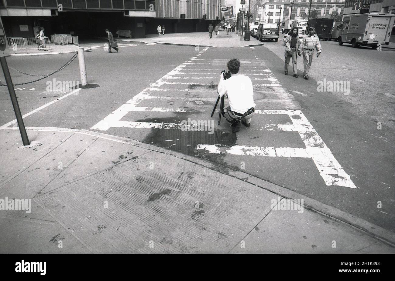 1980, historischer Straßenfotograf, der mit seiner Kamera auf einem kleinen Stativ auf den Straßen von Manahttan in der Mitte der Stadt kniete, in der Nähe des Fußes und des Eingangs zur Tiefgarage von One Penn Plaza, einem 57-stöckigen Wolkenkratzer in New York City, NY, USA. Stockfoto