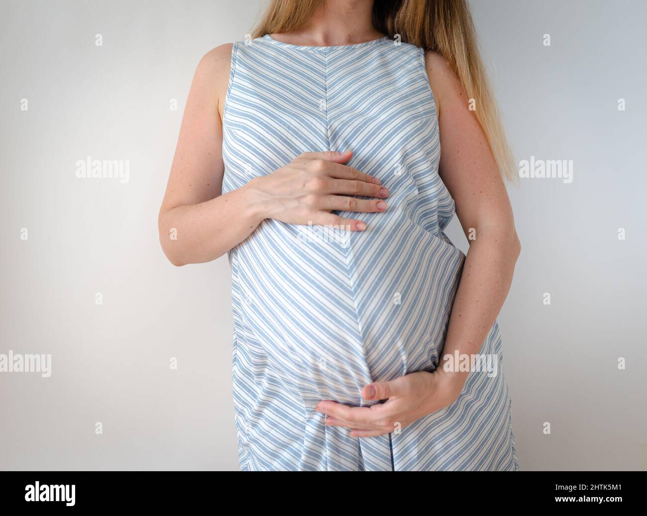 Eine Schwangere in einem blauen Kleid mit weißem Streifen, steht auf weißem Hintergrund und hält ihre Hände auf dem Bauch, das Konzept der Erwartung eines Stockfoto