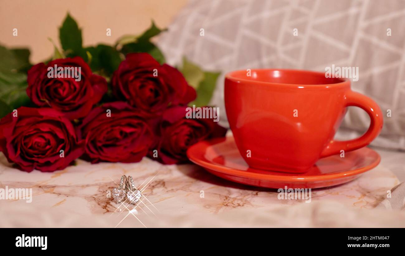 Hintergrund Festliches Flatlay, Komposition zum Valentinstag am 14. Februar. Kaffee in einer roten Tasse, rote Rosenblüten und Diamantherz-Ohrringe zu Hause im Bett Stockfoto