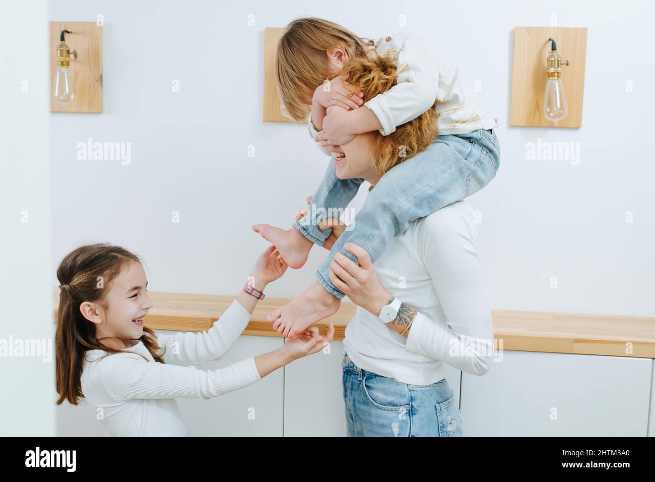 Lachende Schwester kitzelt den kleinen Jungen auf den Füßen, er sitzt auf den Schultern seiner Mutter. Im Flur. Er bedeckt die Augen seiner Mutter mit seinen Händen. Stockfoto