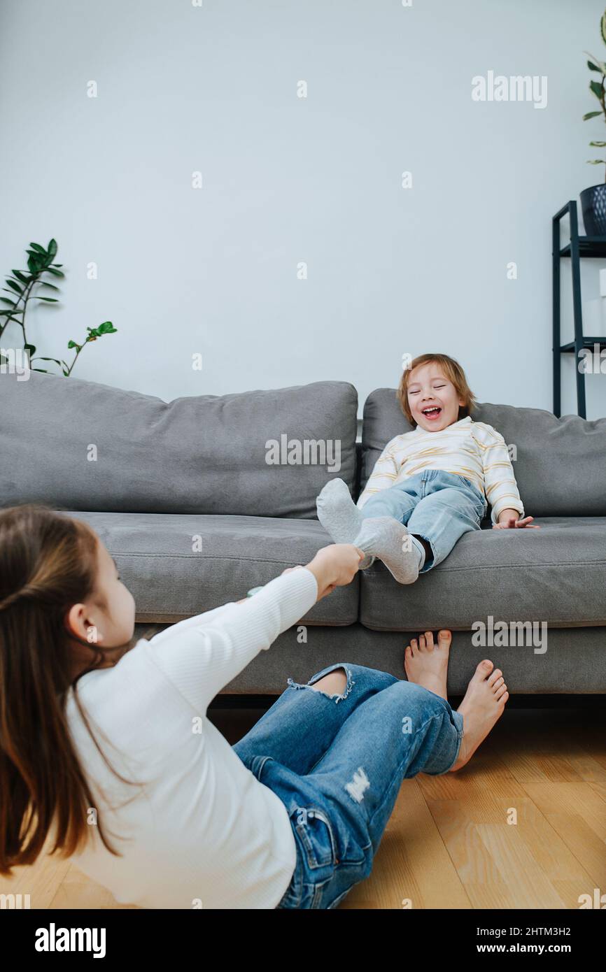 Ein starkes Mädchen, das sich körperlich anstrengt und die Socken eines Jungen auszieht, bereitet sich auf einen kitzelnden Wettkampf vor. Er sitzt auf einer Couch in einem lebenden r Stockfoto