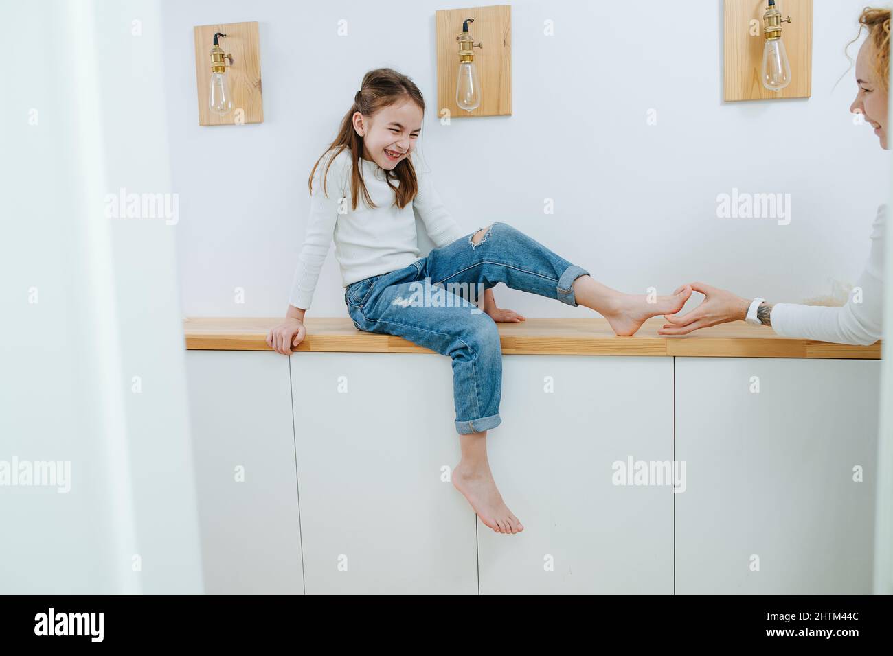 Knirschendes Mädchen, das auf einem Regal im Korridor sitzt und ihre Füße von ihrer Mutter gekitzelt werden. Angespannt und Augen geschlossen. Stockfoto