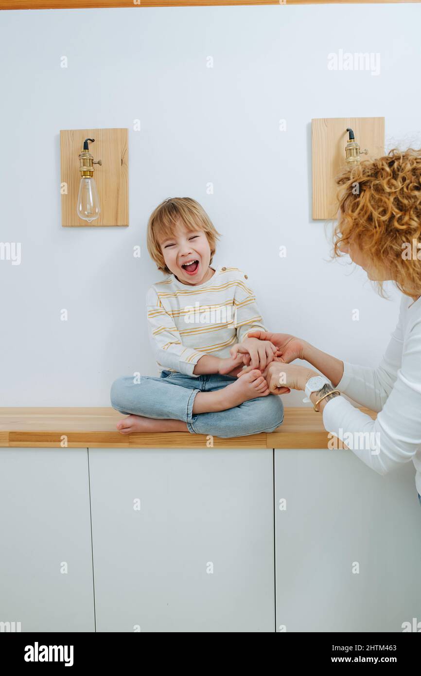 Emotionaler kleiner Junge, der meine Mutter kitzelte. Sitzen mit gekreuzten Beinen auf einem Regal im Flur Stockfoto