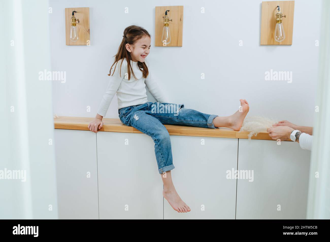 Das Mädchen sitzt auf einem Regal im Korridor und ihre Füße werden von ihrer Mutter gekitzelt. Angespannt und Augen geschlossen. Stockfoto