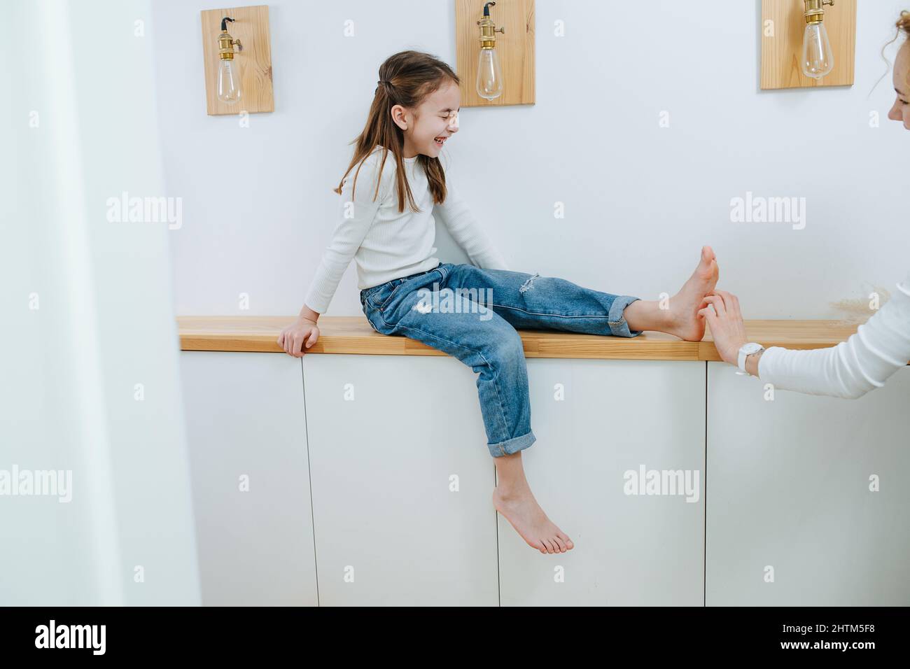 Angespanntes Mädchen, das auf einem Regal im Korridor sitzt und ihre Füße von ihrer Mutter gekitzelt werden. Craining, Augen geschlossen. Stockfoto