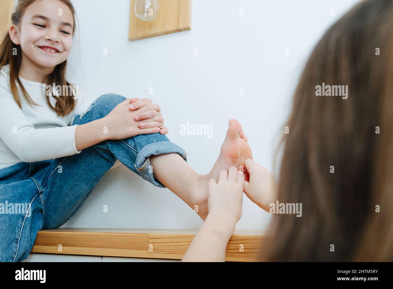 Selbstbewusstes Tween-Mädchen, das auf einem Korridor-Regal sitzt und ihren Fuß von ihrer Schwester kitzelt, sich zeigt und mutig ist. Spaß haben. Stockfoto