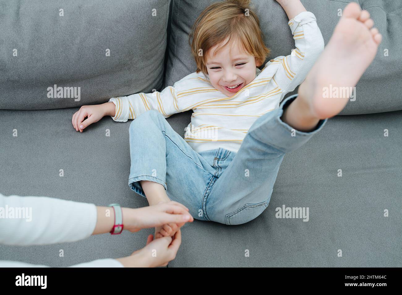 Spritzender lachender Junge, der von den Händen des Mädchens mit dem Fuß auf der Couch gekitzelt wird. Stockfoto