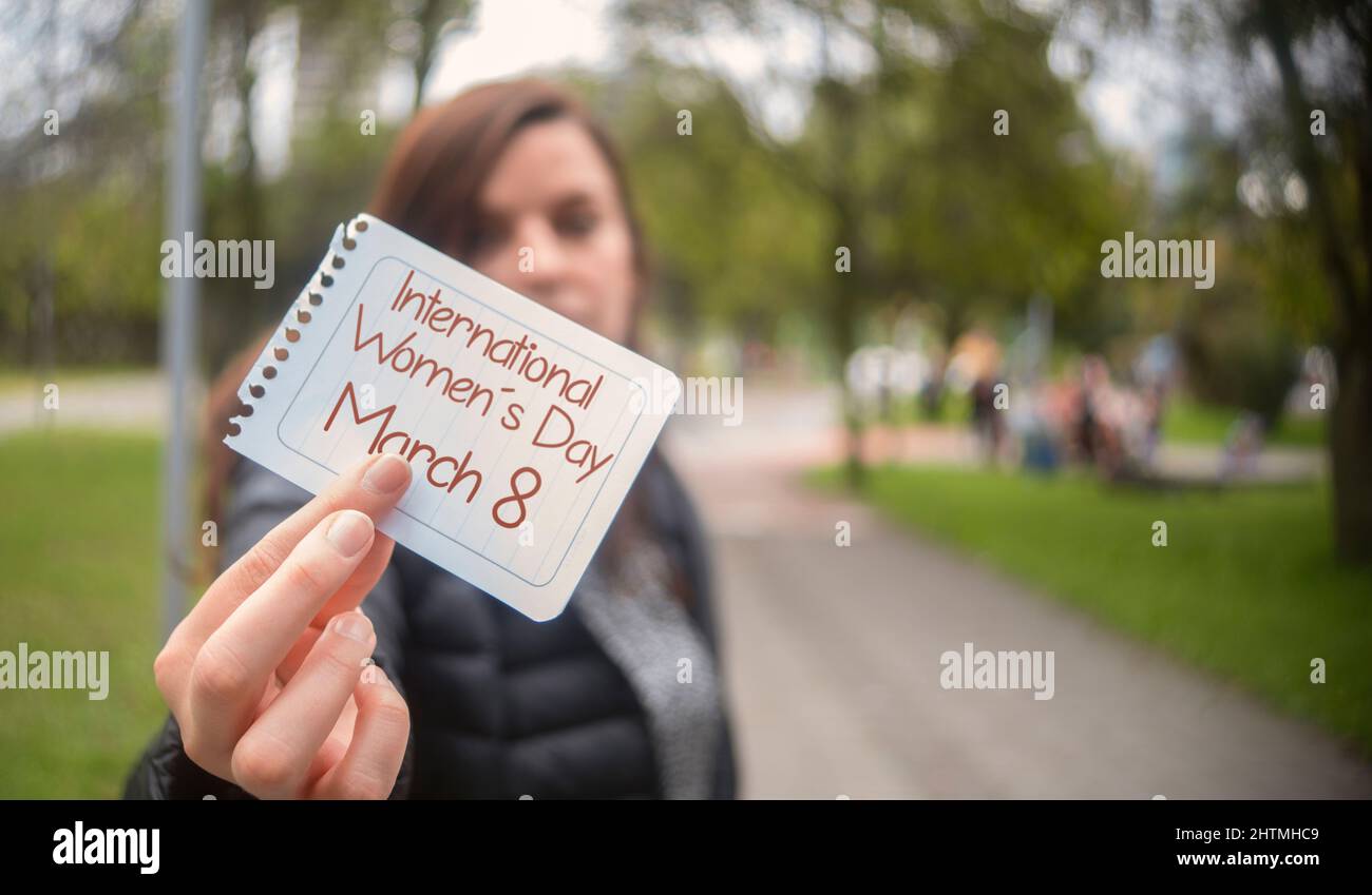 Schöne hispanische Frau mitten in einem Park, die in der Hand ein Papier mit der Botschaft Internationaler Frauentag im Vordergrund hält Stockfoto