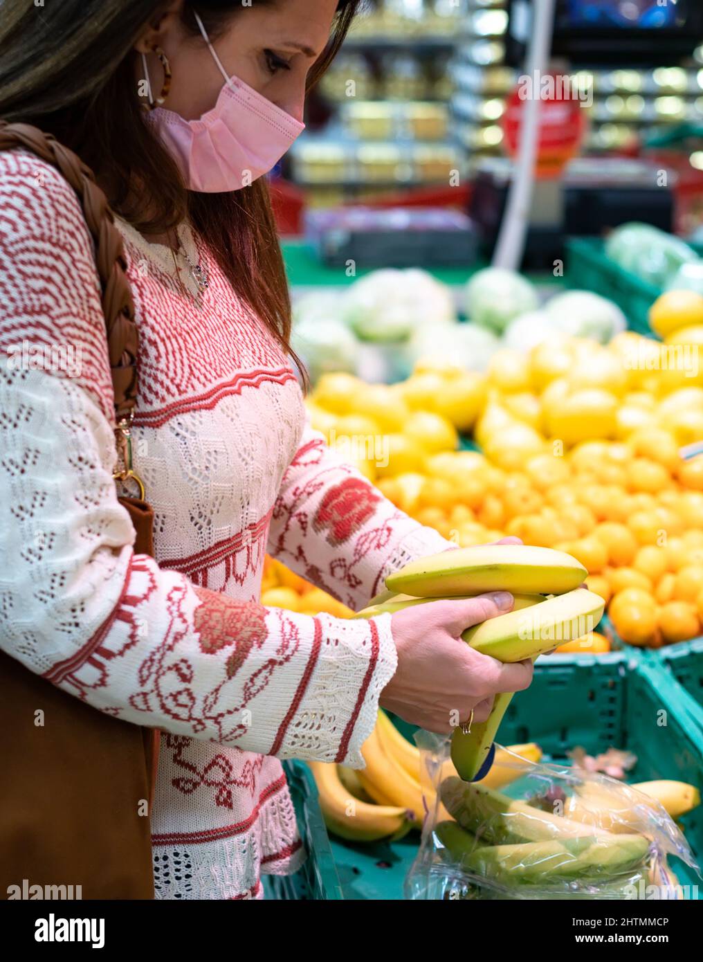 Frau mittleren Alters mit einer rosa Gesichtsmaske hält Bananen in ihren Händen in einem Supermarkt. Sie untersucht die Bananen, um die besten zu kaufen. Stockfoto