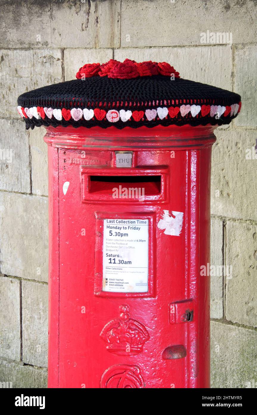 Valentinstag: Ein skurriler, farbenfroher handgefertigter, gehäkelter Wollhut ziert eine traditionelle britische rote Säulenbox. Dorchester, Dorset, England. Stockfoto