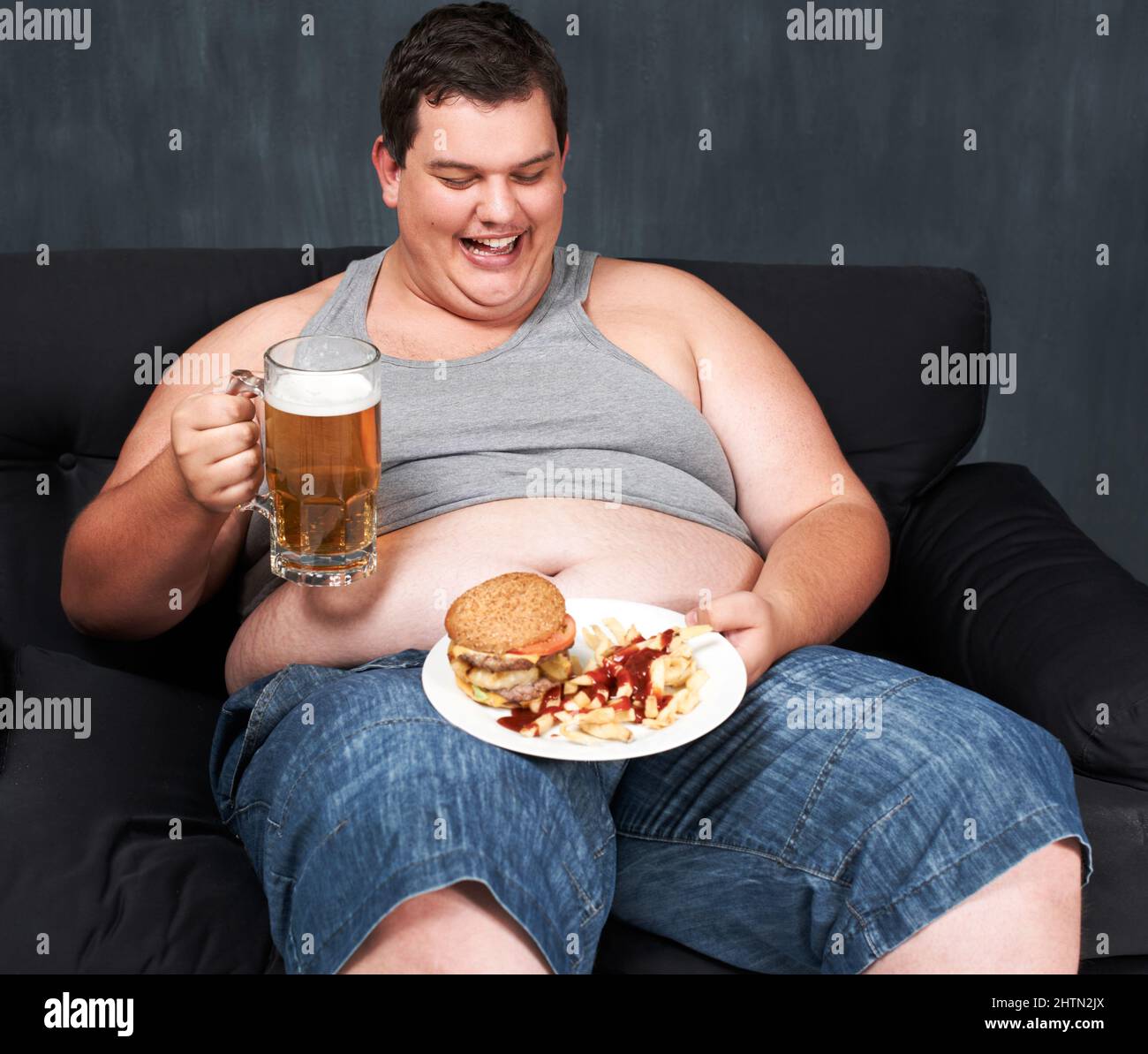Etwas, um sein Essen abzuwaschen. Ein übergewichtiger junger Mann, der auf einem Sofa sitzt und sich an Bier und Fast Food trinkt. Stockfoto