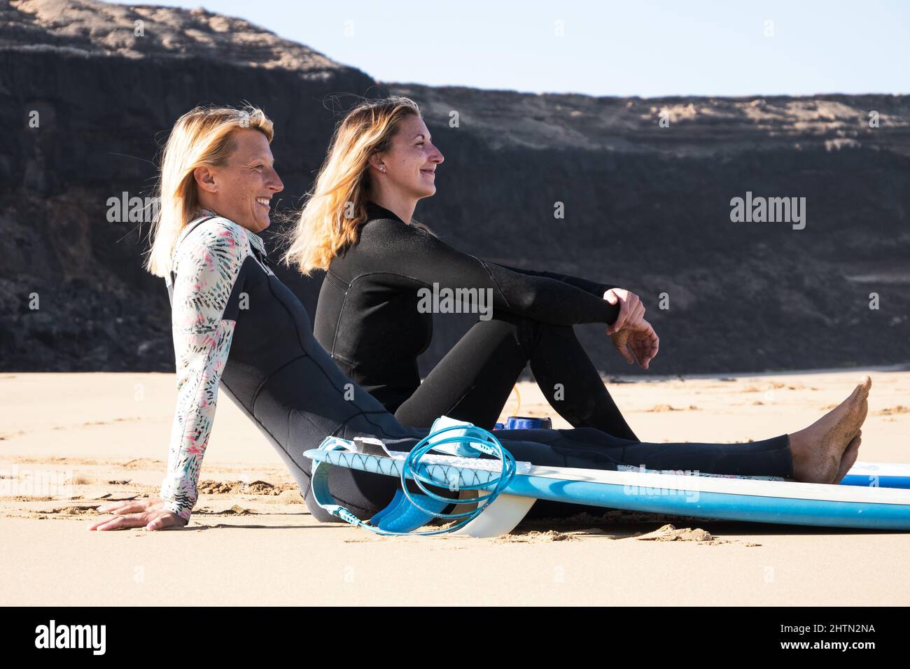 2 kaukasische Surfer sitzen am Strand. Sie lächeln zum Meer. Sport, gesunder Lebensstil, Freundschaftskonzept. Stockfoto