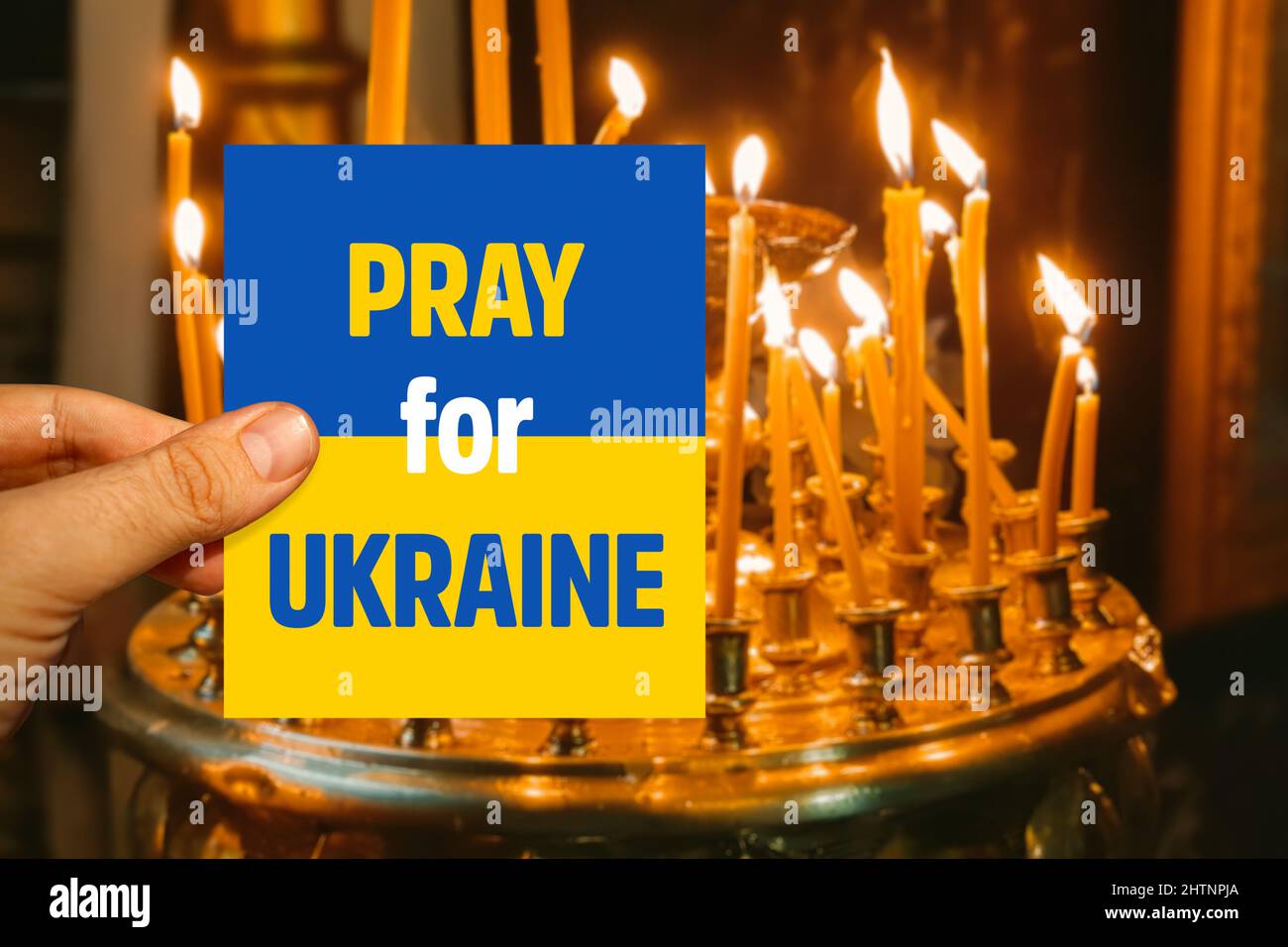 Männliche Hand mit Ukraine-Flaggenkarte und Gebetsbotschaft für die Ukraine vor brennenden Kerzen in einer Kirche in Kiew Stockfoto