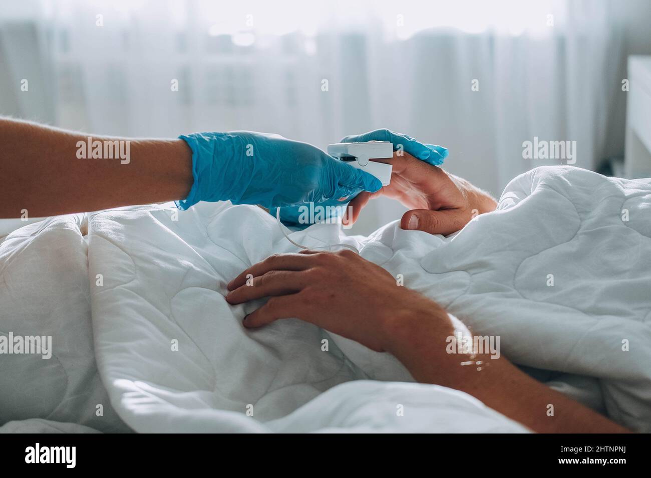 Der Mann liegt im Bett, ein Arzt mit medizinischen Handschuhen kommt auf ihn zu und legt ein Pulsoximeter auf seinen Finger, um die Sauerstoffsättigung zu messen. Asthmabehandlung Stockfoto