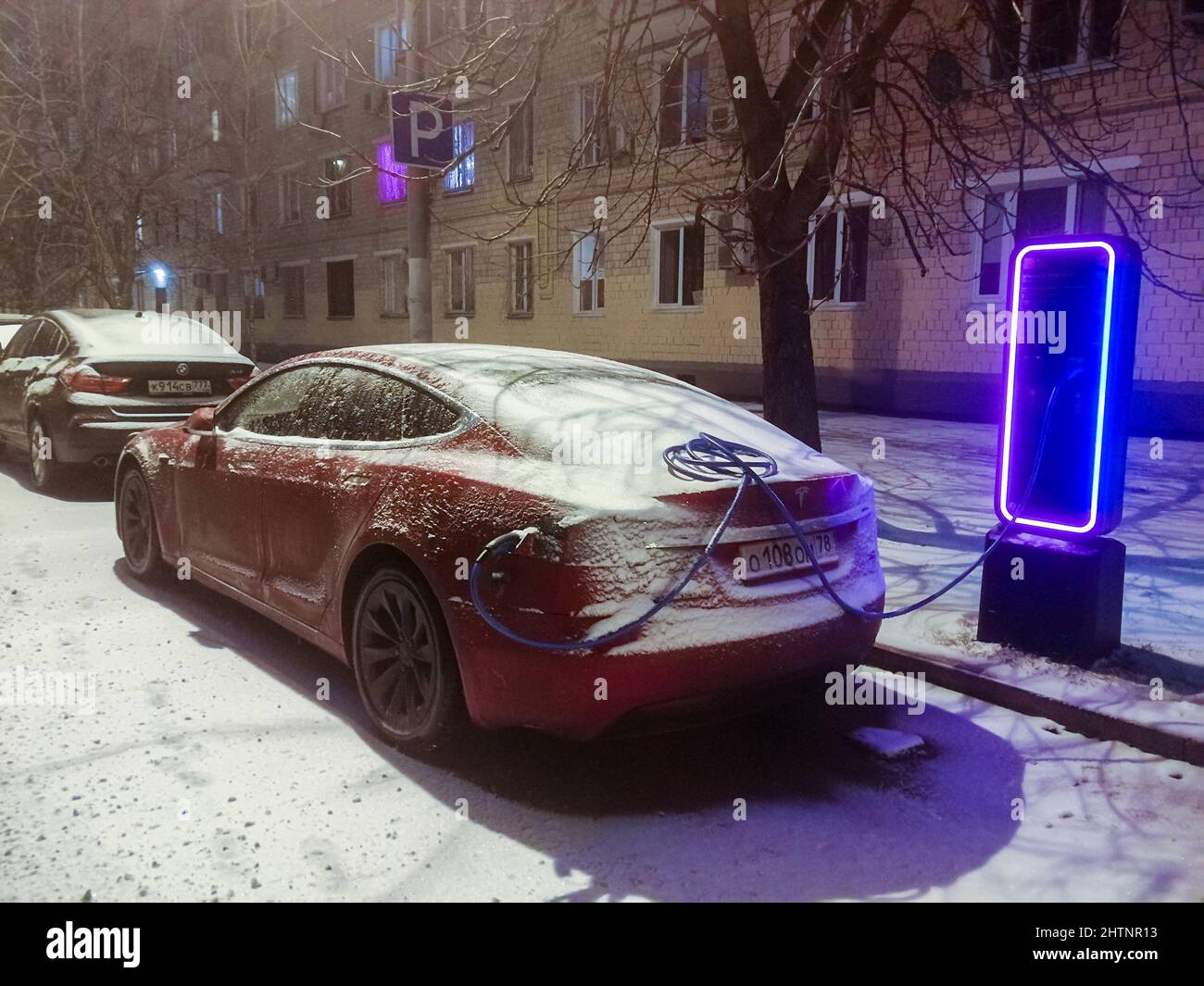 Moskau. Russland. 30. November 2021. Ein Tesla-Auto lädt in einer Winternacht an einer Ladestation auf einer Stadtstraße eine Batterie auf. Ein rotes, emissionsfreies Tesla-Auto lädt auf einer Stadtstraße. Stockfoto
