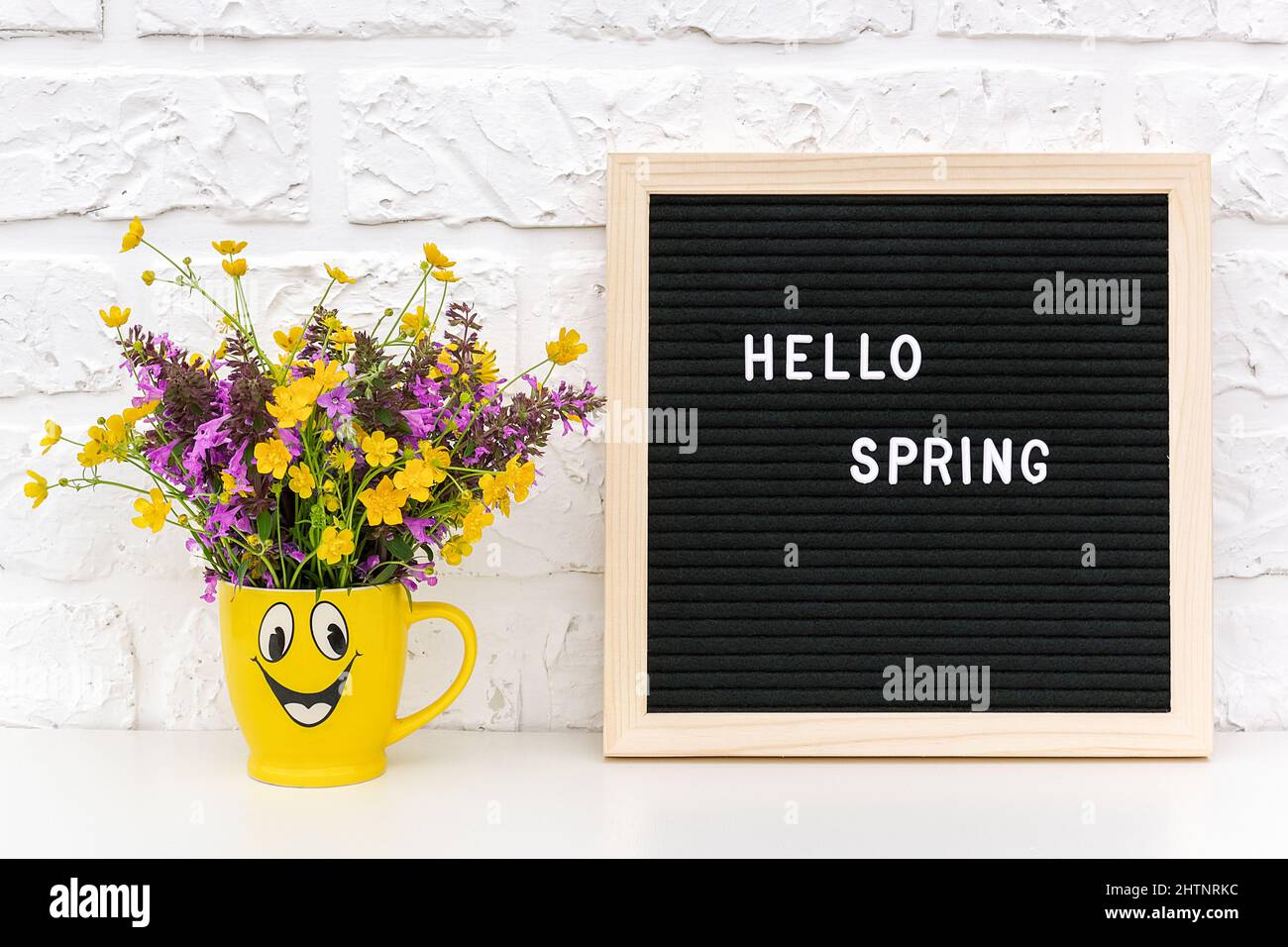 Text Hallo Frühling auf schwarzem Briefbrett und Blumenstrauß in lustiger gelber Tasse auf weißem Backsteinwand. Konzept Springtime Stimmung und Stockfoto