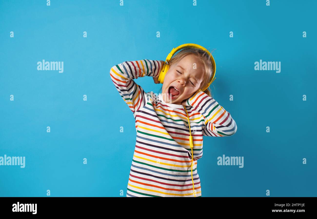 Porträt eines niedlichen, charmanten Mädchens, das ihr Lieblingslied in gelben Kopfhörern lautstark mit offenem Mund auf blauem Hintergrund singt Stockfoto