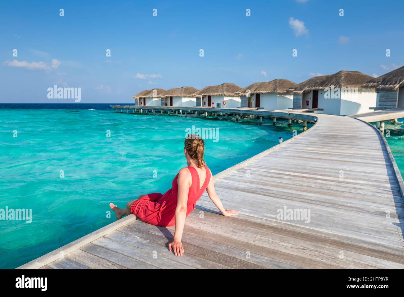 Strandurlaub mit Frau, die sich auf einem hölzernen Pier im luxuriösen Hotelresort auf den Malediven mit Überwasser-Villen, türkisfarbenem Meerwasser und blauen s entspannt Stockfoto