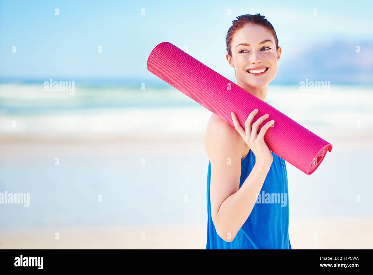 Auf der Suche nach einem guten Ort, um etwas Yoga zu machen. Aufnahme einer jungen Frau, die eine Yogamatte in der Hand hält, während sie am Strand steht. Stockfoto