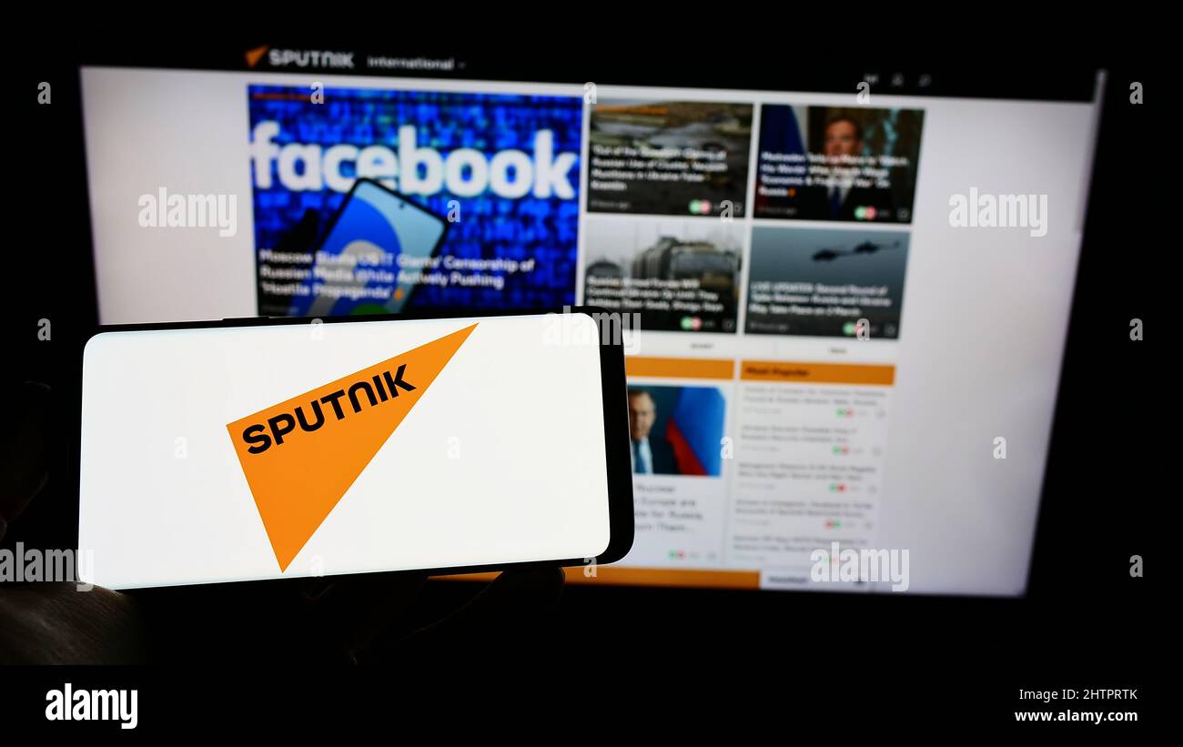 Person, die Mobiltelefon mit dem Logo des russischen staatlichen Medienunternehmens Sputnik auf dem Bildschirm vor der Webseite hält. Konzentrieren Sie sich auf die Telefonanzeige. Stockfoto