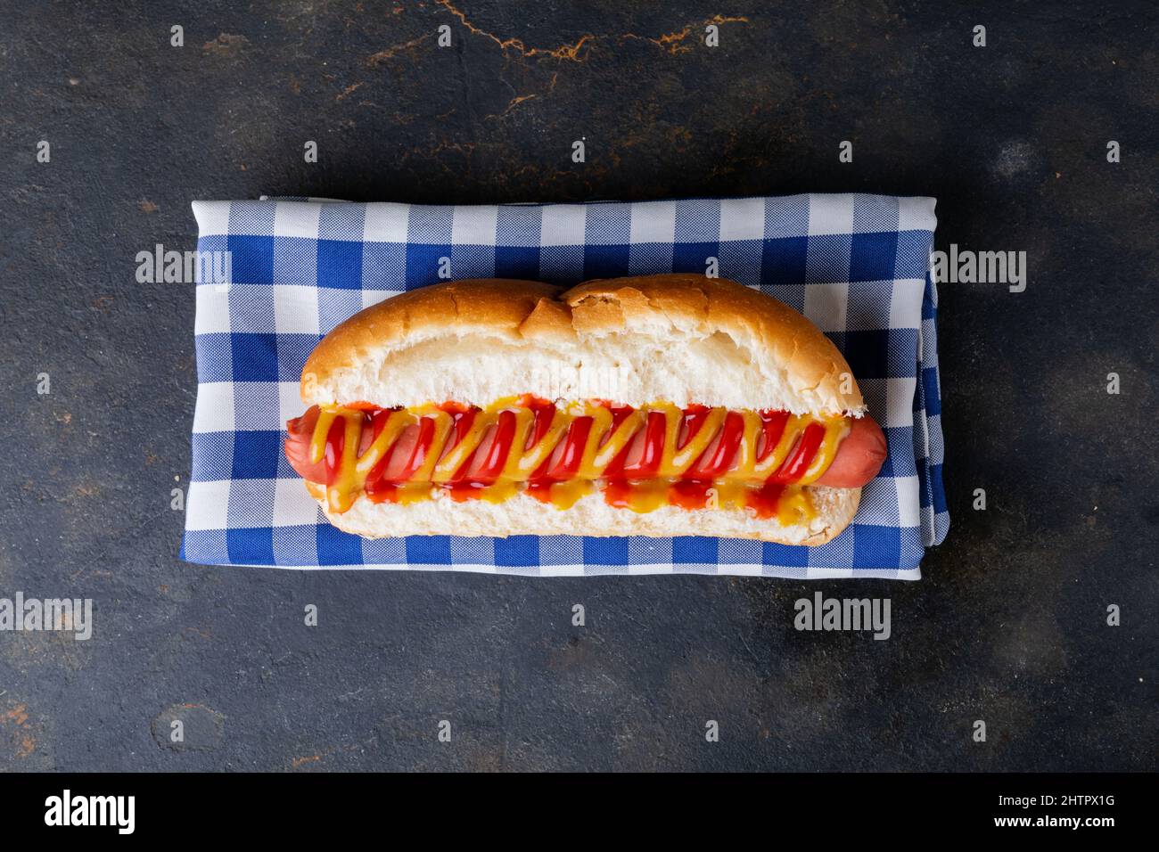 Blick von oben auf Hot Dog mit Tomate und musterter Sauce auf karierter Serviette am Tisch Stockfoto
