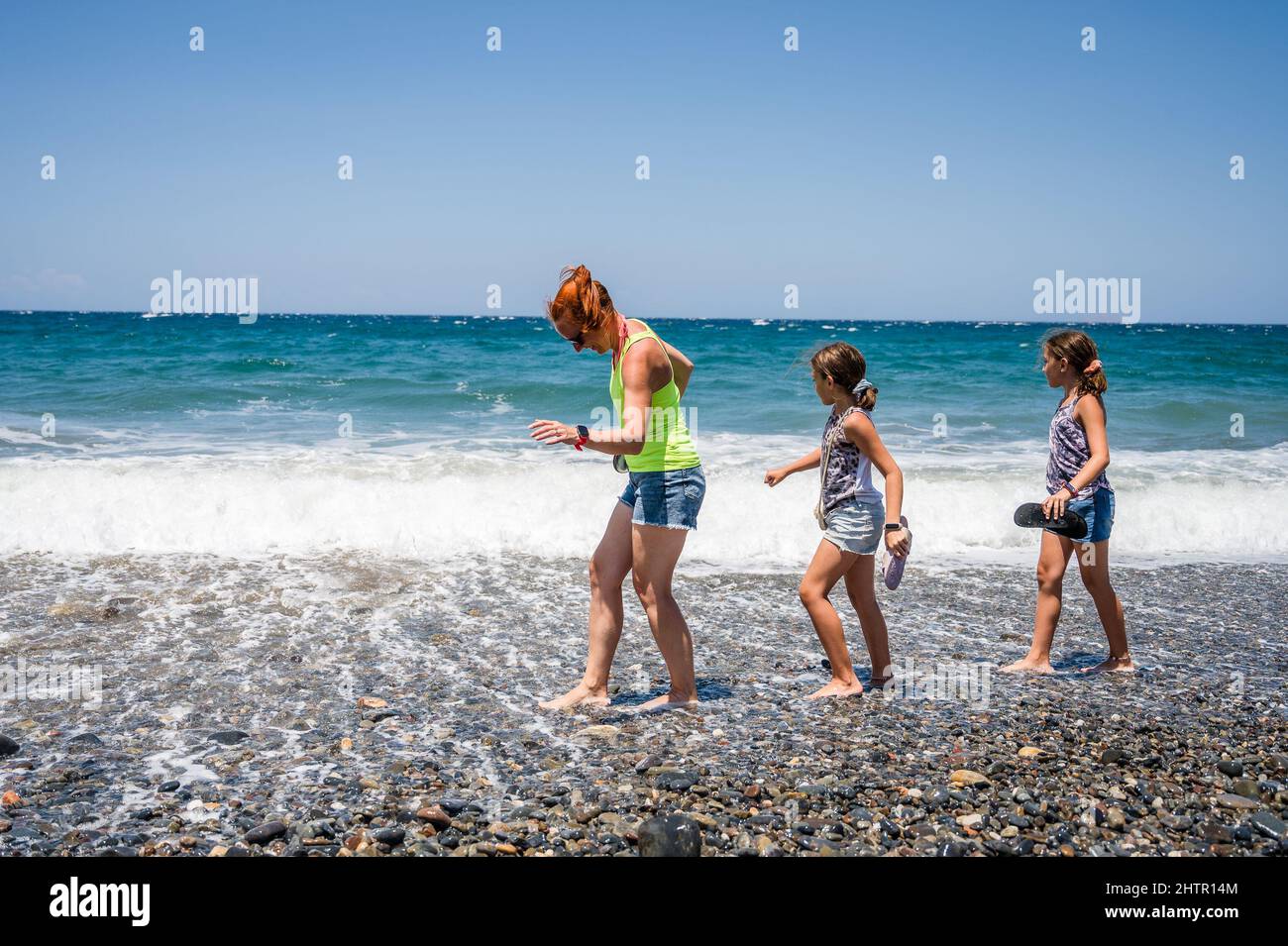 Mutter und Töchter gehen auf einem aktiven Familienurlaub am Strand spazieren. Familienwanderungen an einem schönen Strand mit kleinen Steinen und Meereswellen. Kreta, Stockfoto