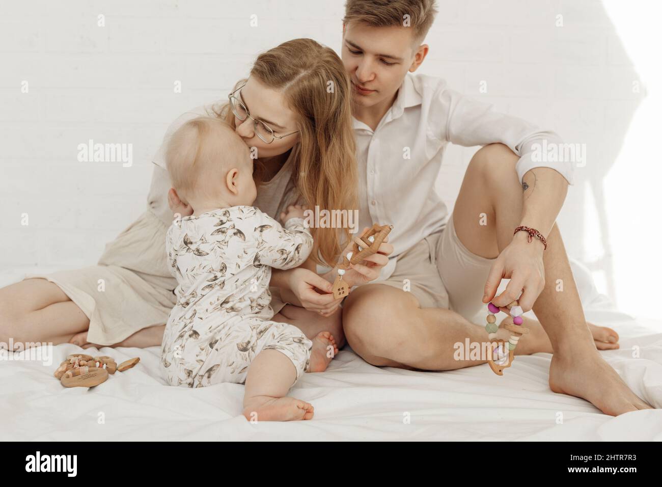 Porträt einer jungen, schönen Familie mit einem dicken Säugling, der einen modernen, umweltfreundlichen Beißring aus Holz auf einem weißen Bett hält. Stockfoto