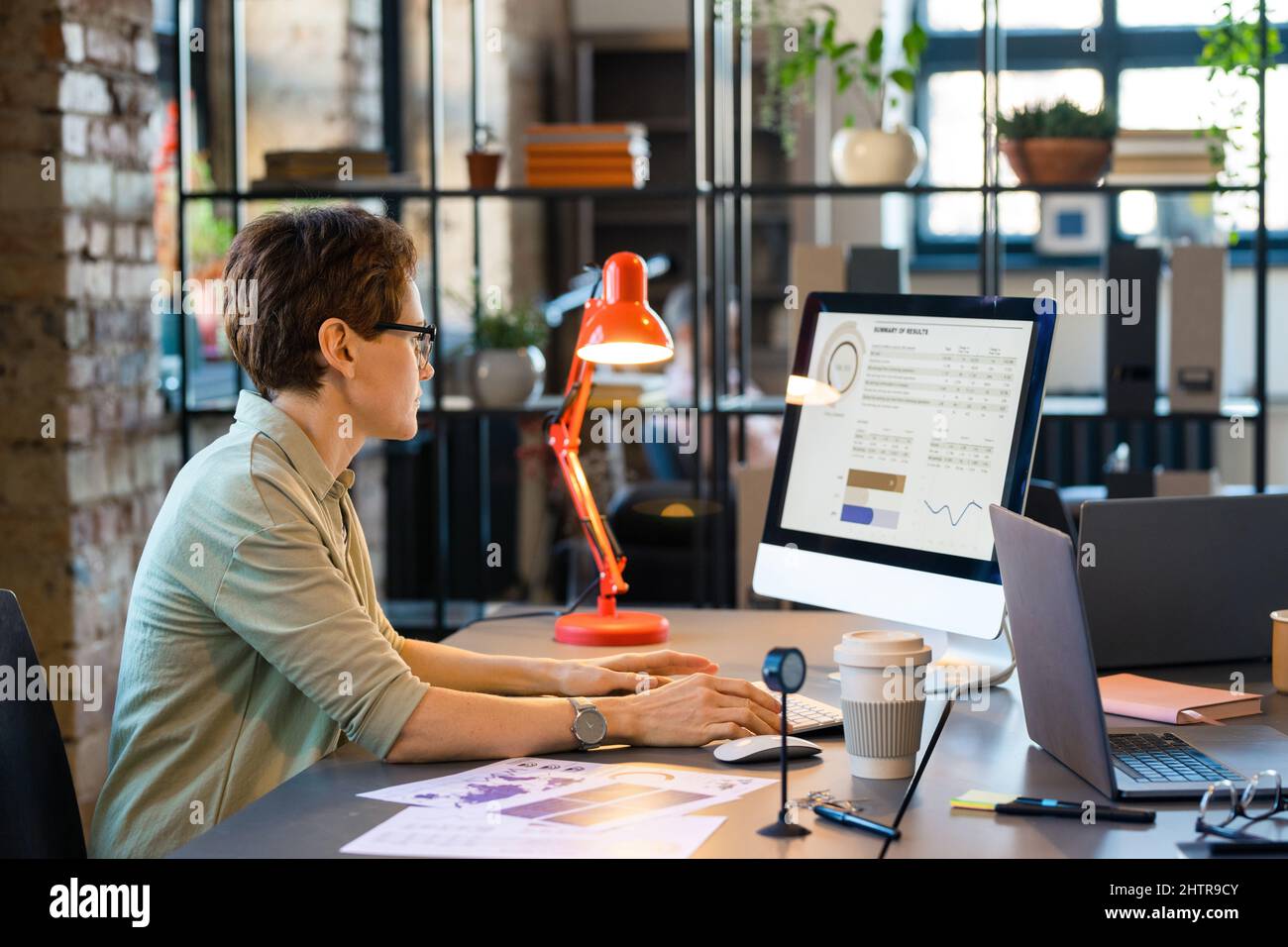 Geschäftsfrau in Brillen konzentriert sich auf ihre Arbeit am Computer, sie sitzt am Tisch und untersucht Grafiken auf dem Monitor Stockfoto