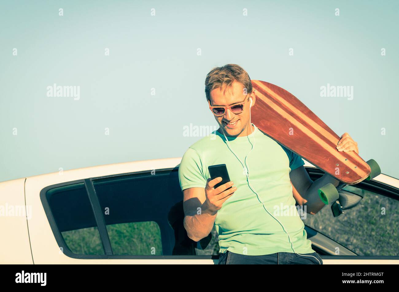 Junger Hipster-Mann mit Smartphone, der Musik während der Ausreispause neben seinem Auto hört - Konzept neuer Trends und moderner Technologien gemischt mit einem Retro-li Stockfoto