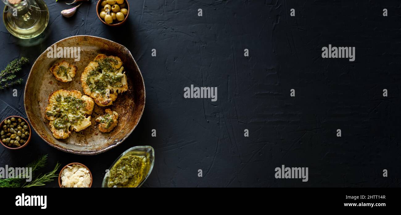 Blumenkohlesteak mit Gewürzen liegt in einer Pfanne. Olivenöl, Chimichurri-Sauce, Kräuter, verschiedene Gewürze nebeneinander. Dunkler Hintergrund. Vegetarische Gerichte Stockfoto