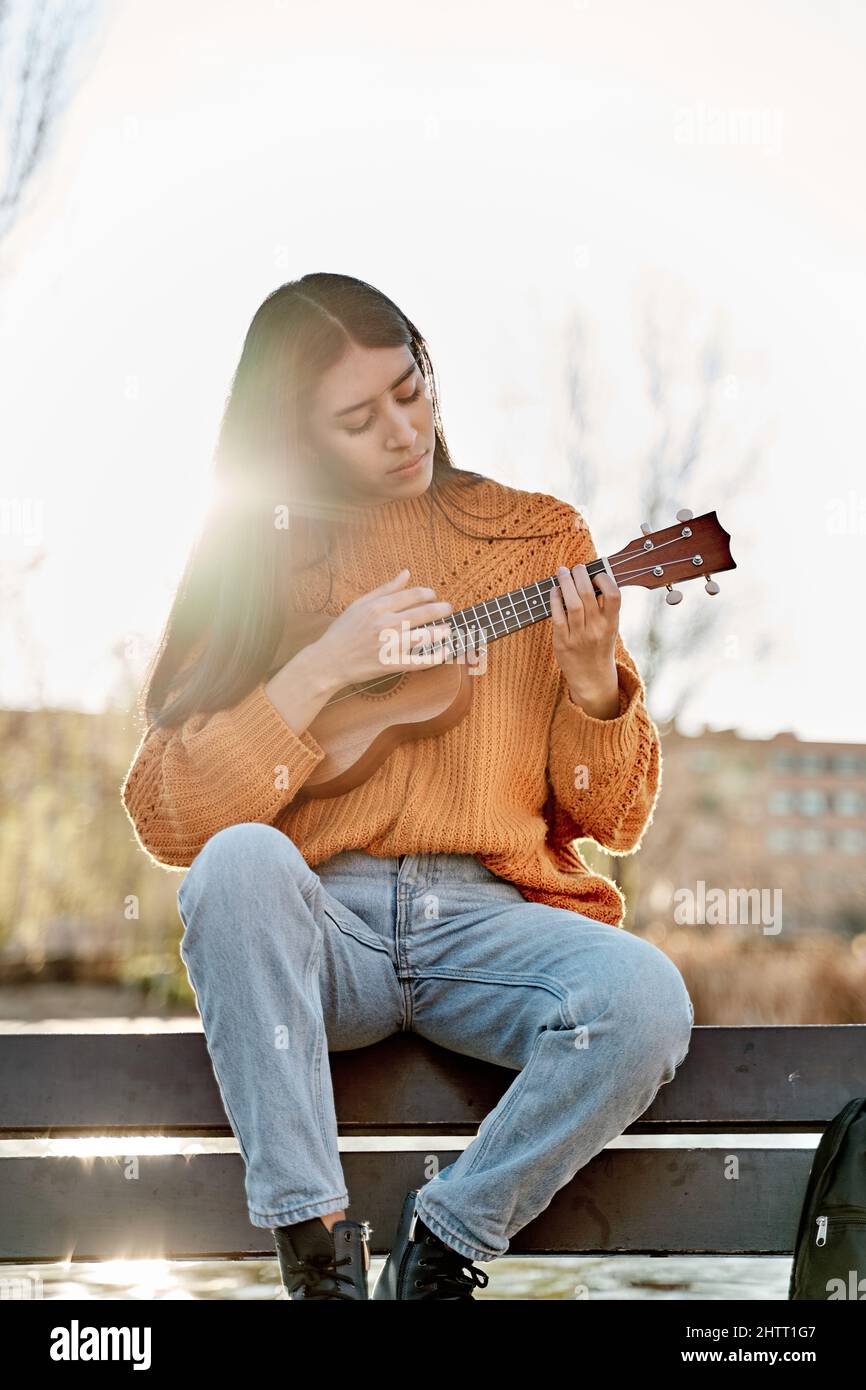 Junge Latina spielt Ukulele in einem Stadtpark. Frau sitzt auf einer Bank und übt mit ihrem Musikinstrument. Stockfoto