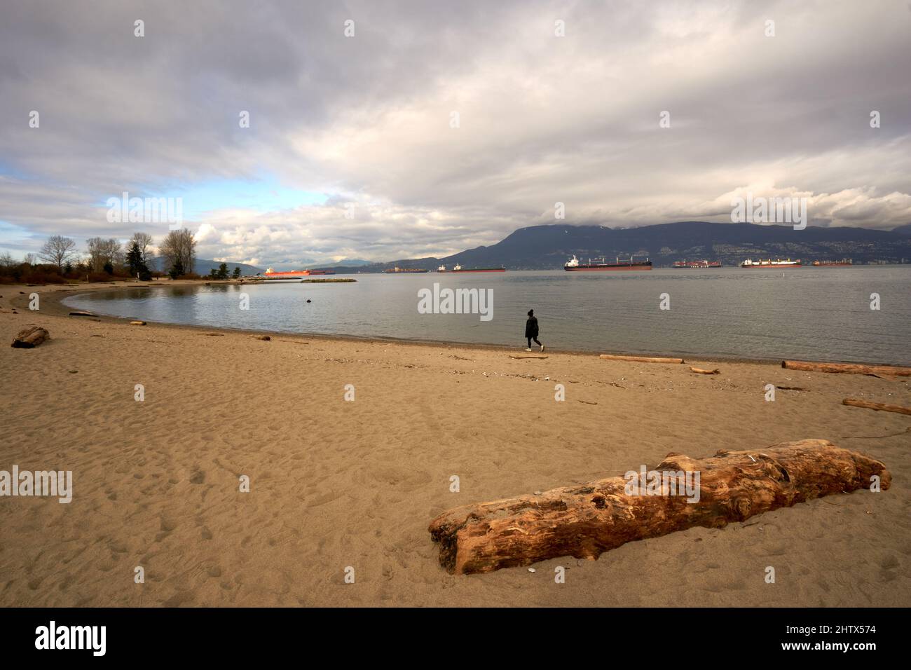 Frau, die im Winter mit der English Bay mit Frachtern im Hintergrund am Strand von New York entlang läuft, Vancouver, British Columbia, Kanada Stockfoto