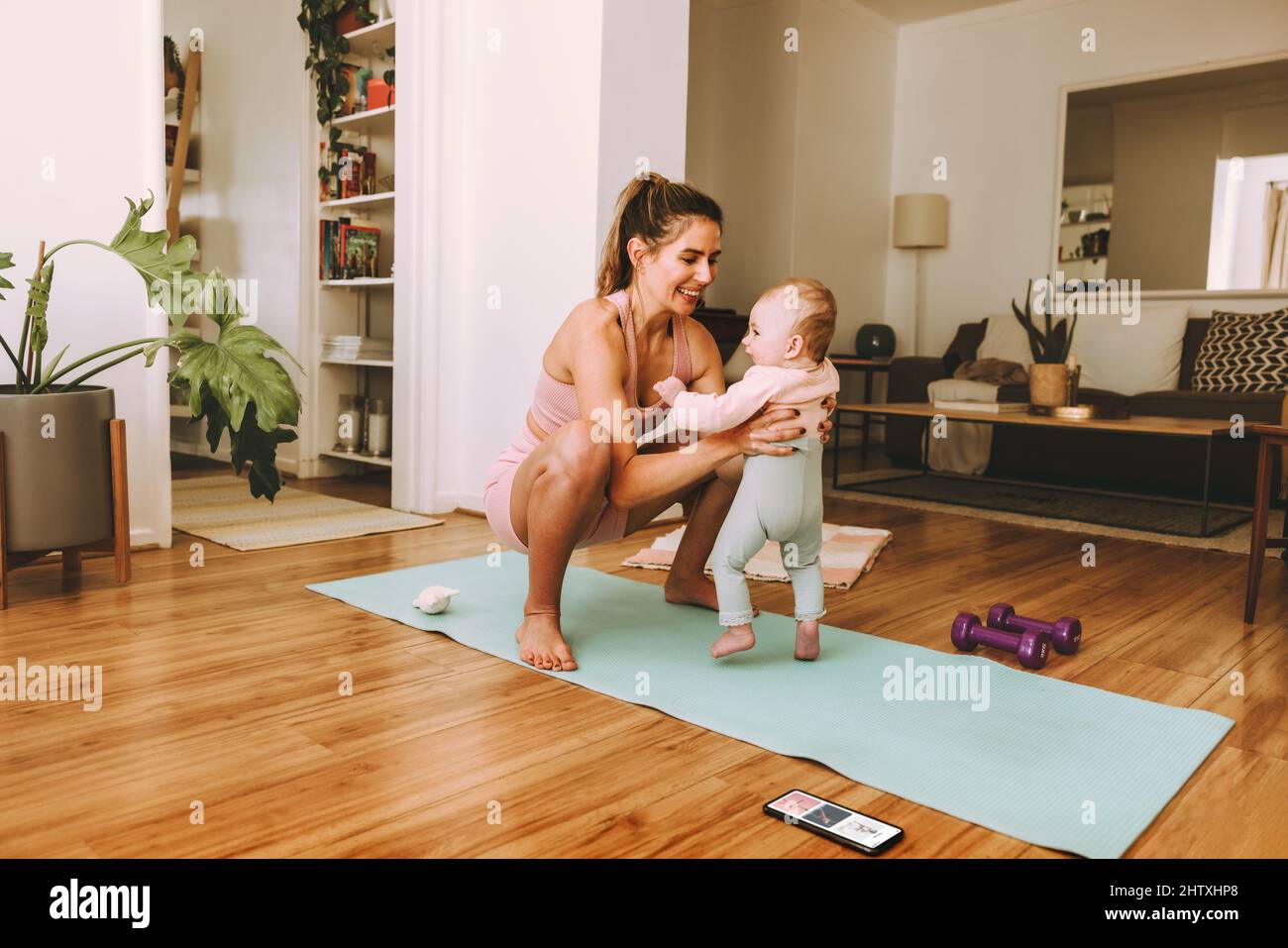 Yogi-Mutter hebt ihr Baby auf einer Übungsmatte. Glückliche Mutter lächelt fröhlich, während sie mit ihrem Baby zu Hause arbeitet. Neue Mutter Bonding mit ihrem Baby d Stockfoto